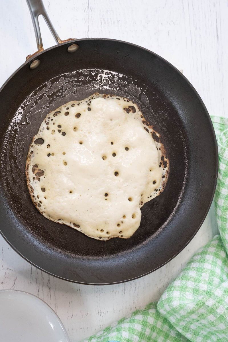 A cast iron skillet holds a cooking pancake with bubbles on top.