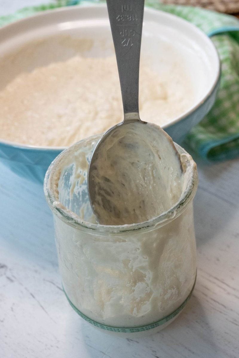 A spoon sits in a coated but empty glass jar next to a mixing bowl with sourdough starter.