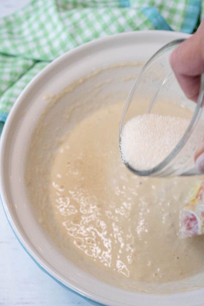 A hand pours dry white ingredients from small glass bowl into bigger bowl of batter.