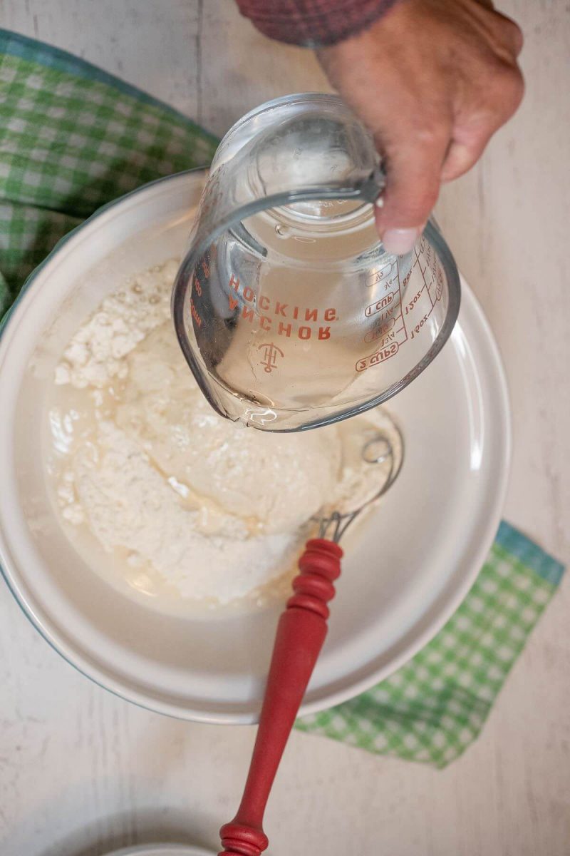 A hand pours a glass measuring cup of water into bowl of sourdough starter.