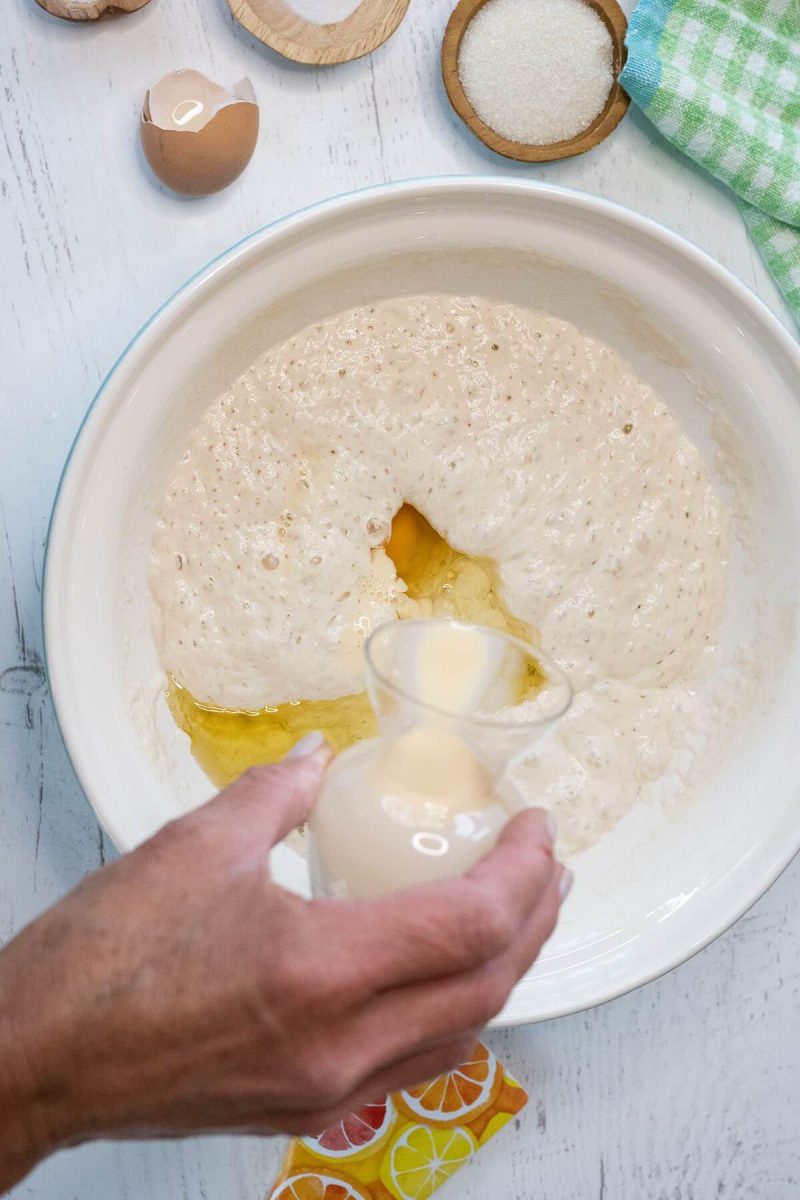 A hand pours a small carafe of oil into the sourdough sponge in a bowl.
