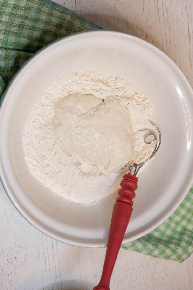 A white bowl holds flour, sourdough starter batter, and a red whisk.