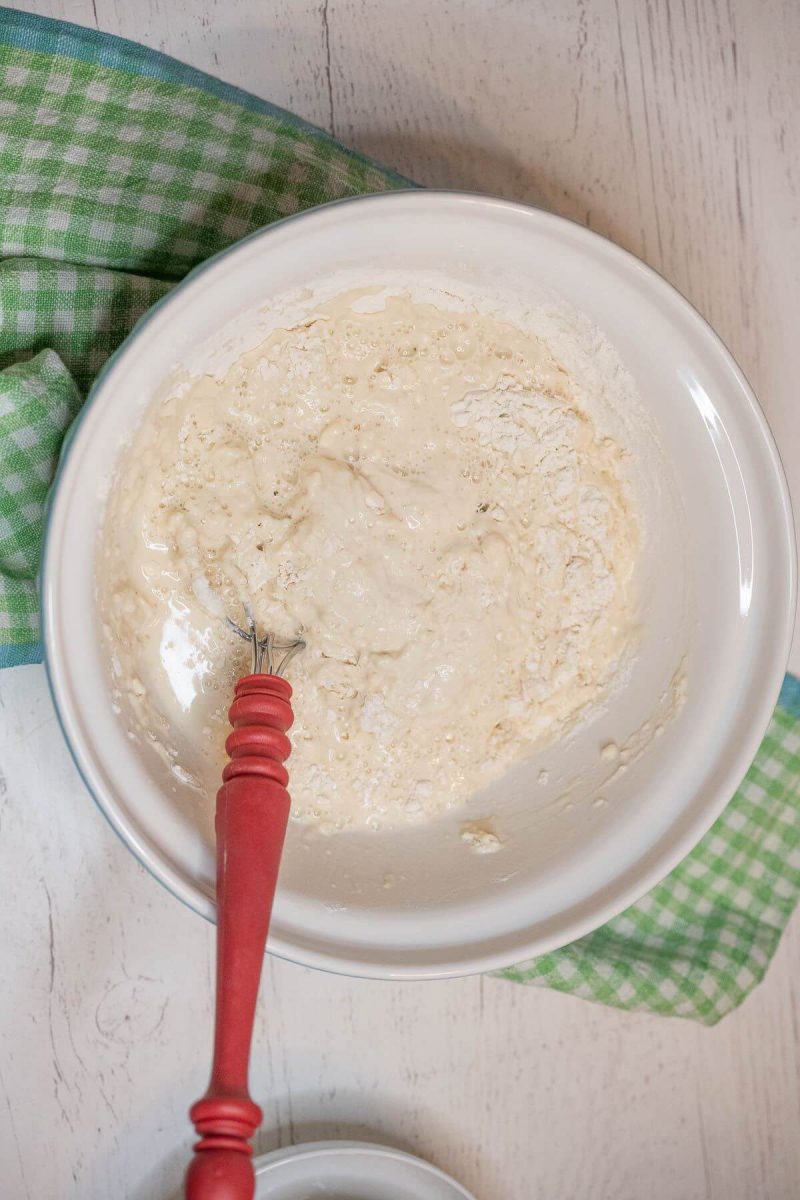 A red whisk stirs flour into sourdough starter batter in bowl.