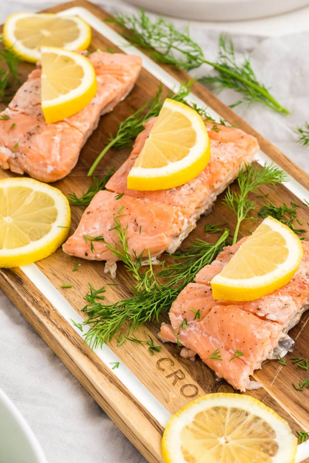 Cooked salmon pieces are topped with lemon slices on cut board with fresh dill.