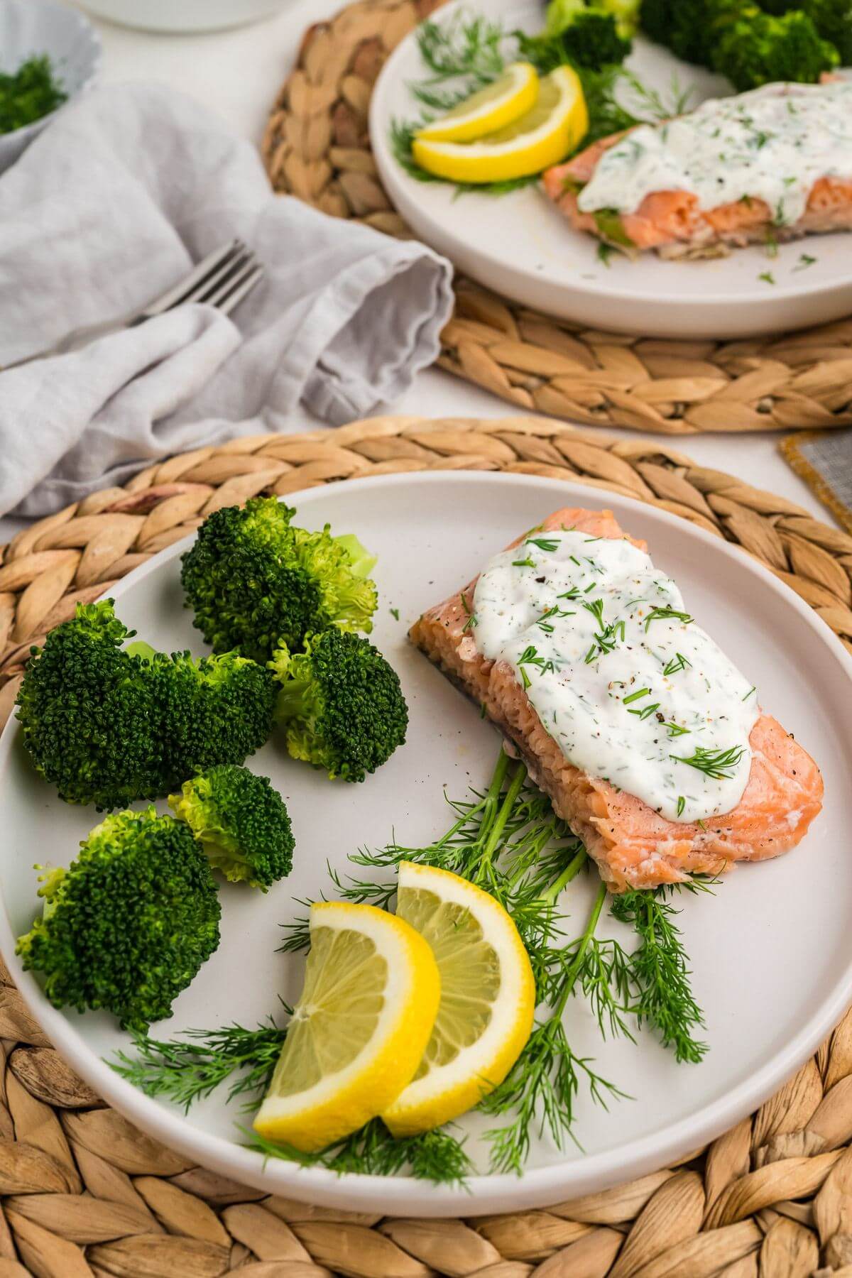 Two plates filled with broccoli, dill, lemon, and salmon with sauce are on a table.