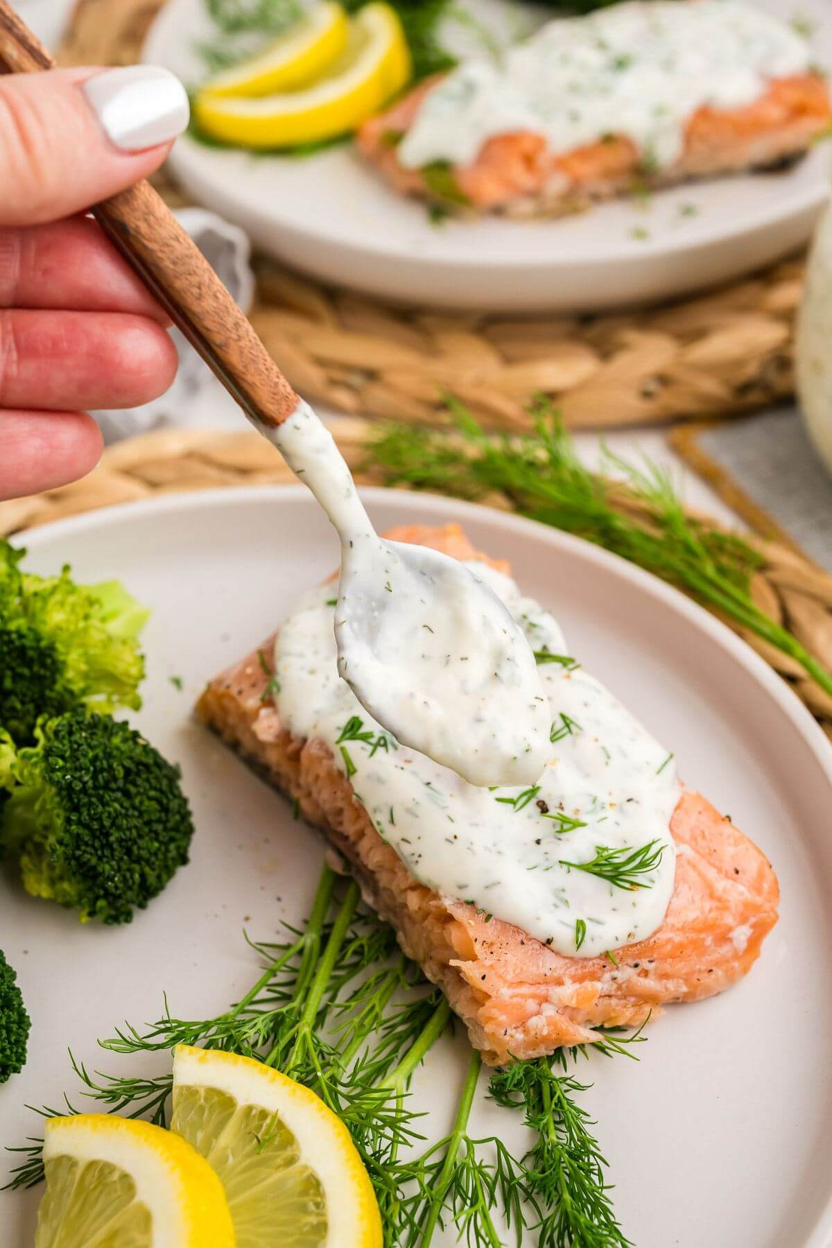 A hand uses a spoon to dollop white sauce on top of salmon.