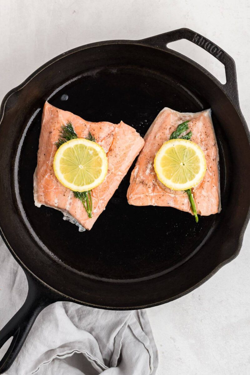Two salmon filets are in an iron skillet and topped with dill and lemon.