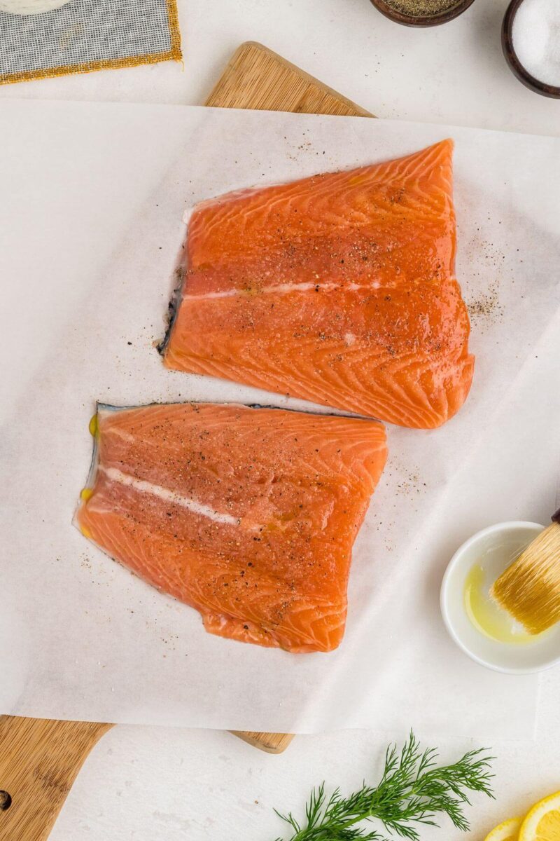 Two uncooked salmon filets rest on parchment paper near a brush with oil.