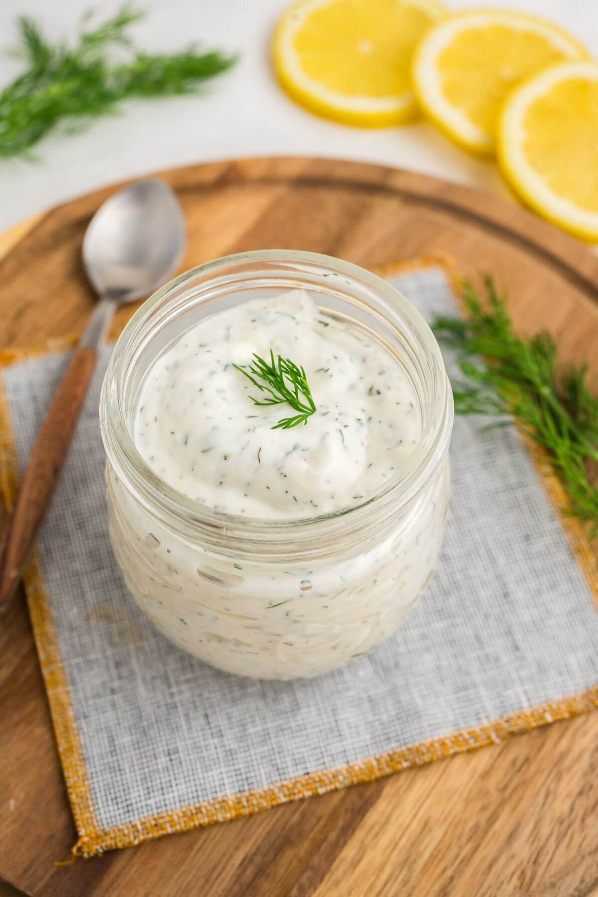 Lemon slices and fresh dill frame a small jar of white sauce with green specks on a serving board.