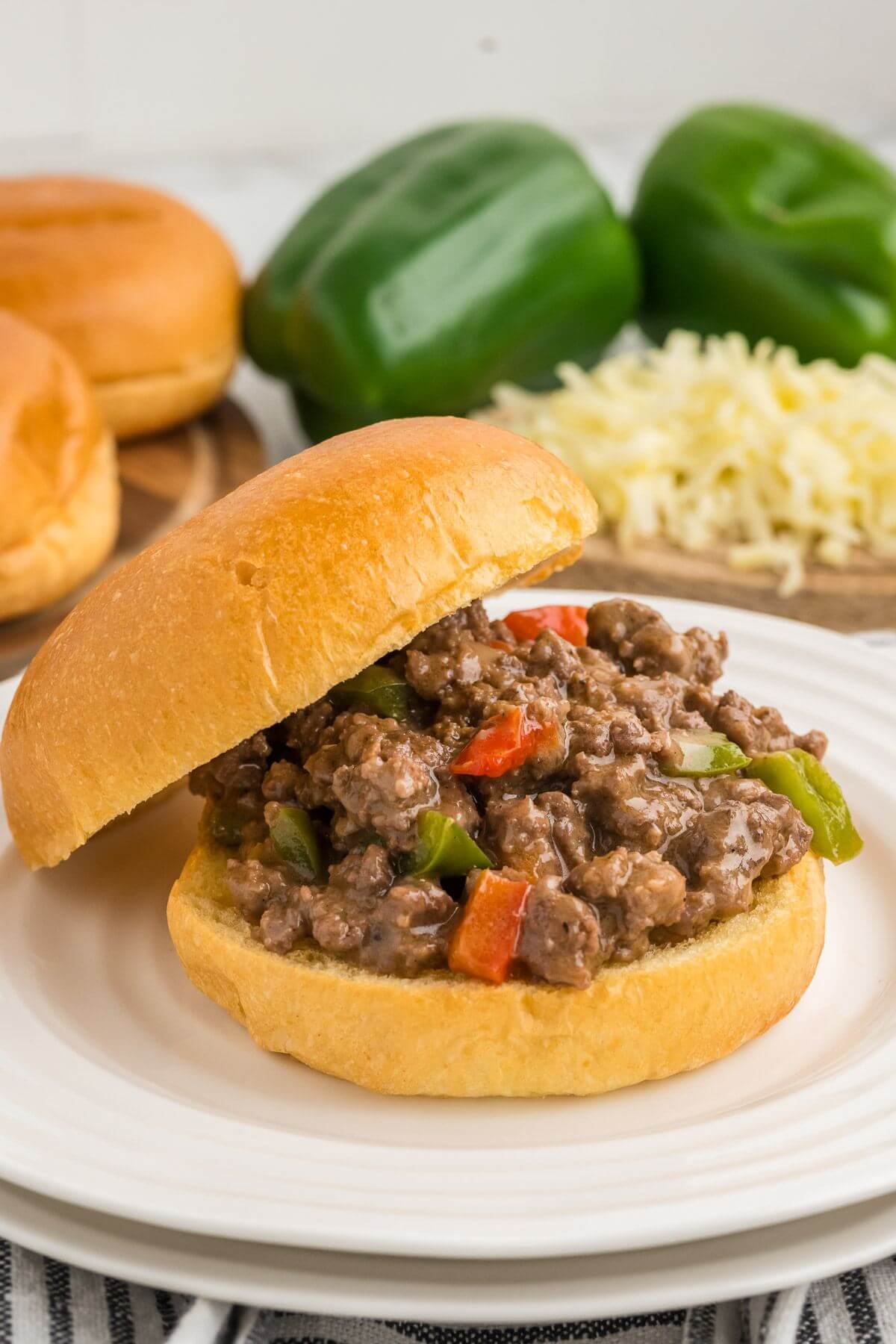Extra bell peppers, cheese, and buns are behind a full and meaty sandwich with its bun tilting back.
