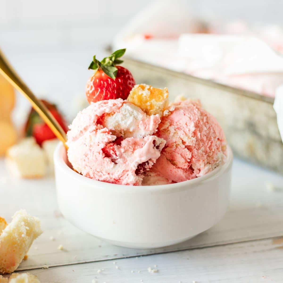 Bowl with three scoops of strawberry shortcake ice cream, with a spoon and a fresh strawberry on top.