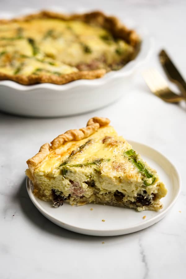 Asparagus and ham quiche with slice on a plate and pie plate in background.