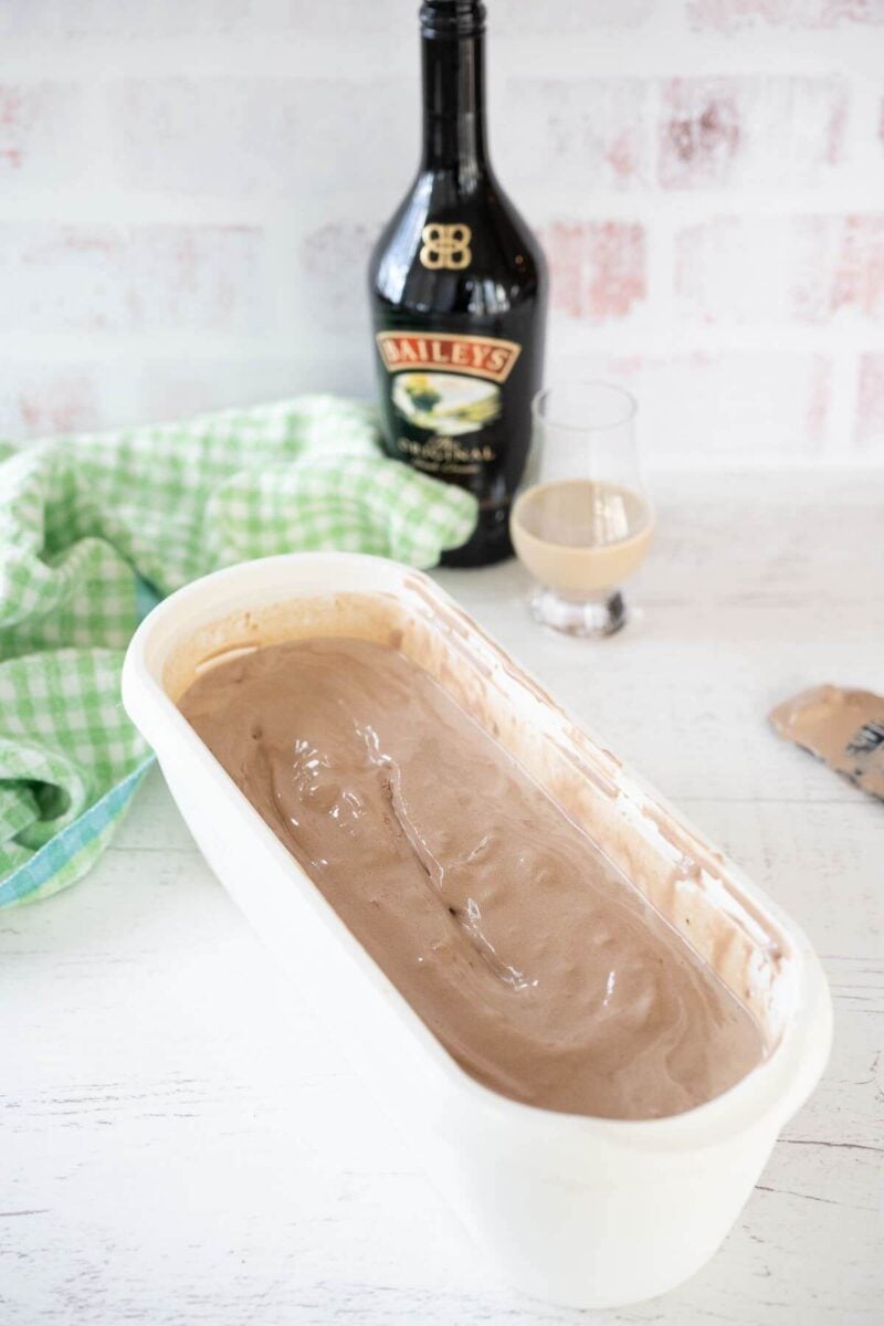 A bottle and glass of Baileys stand behind a long pan filled with soft serve ice cream.