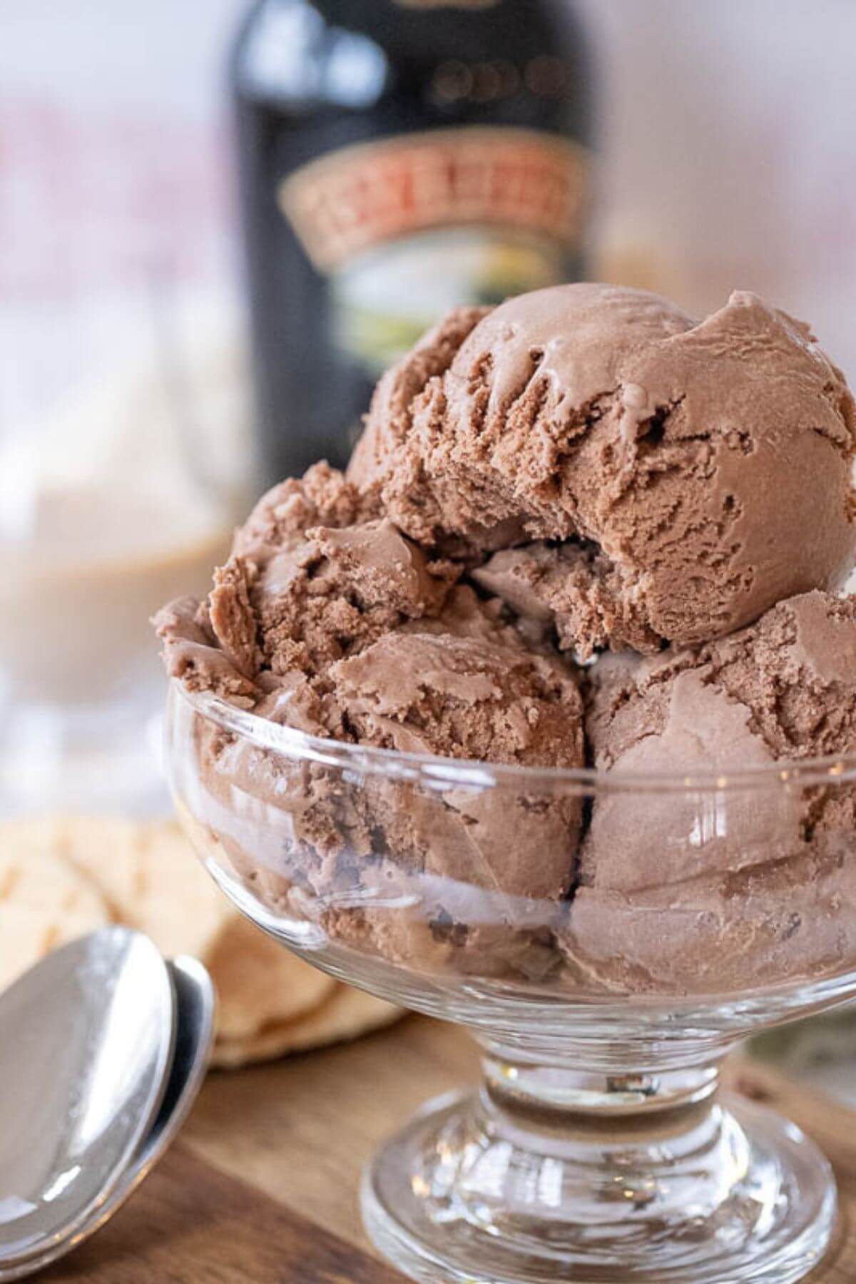 A close up shows the creamy texture of ice cream scoops filling a glass bowl with bottle of Baileys behind it.