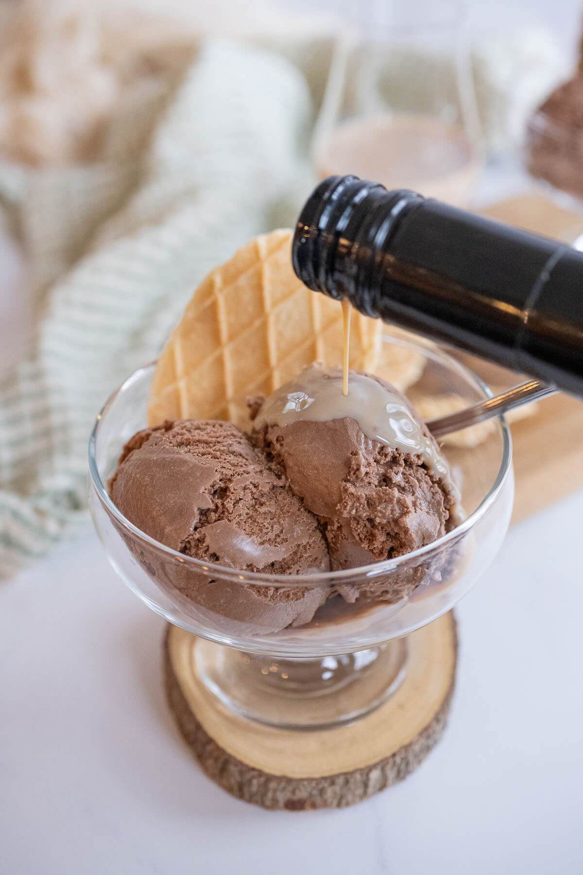 A dark bottle neck pours out creamy liquor over scoops of ice cream in a clear dish.