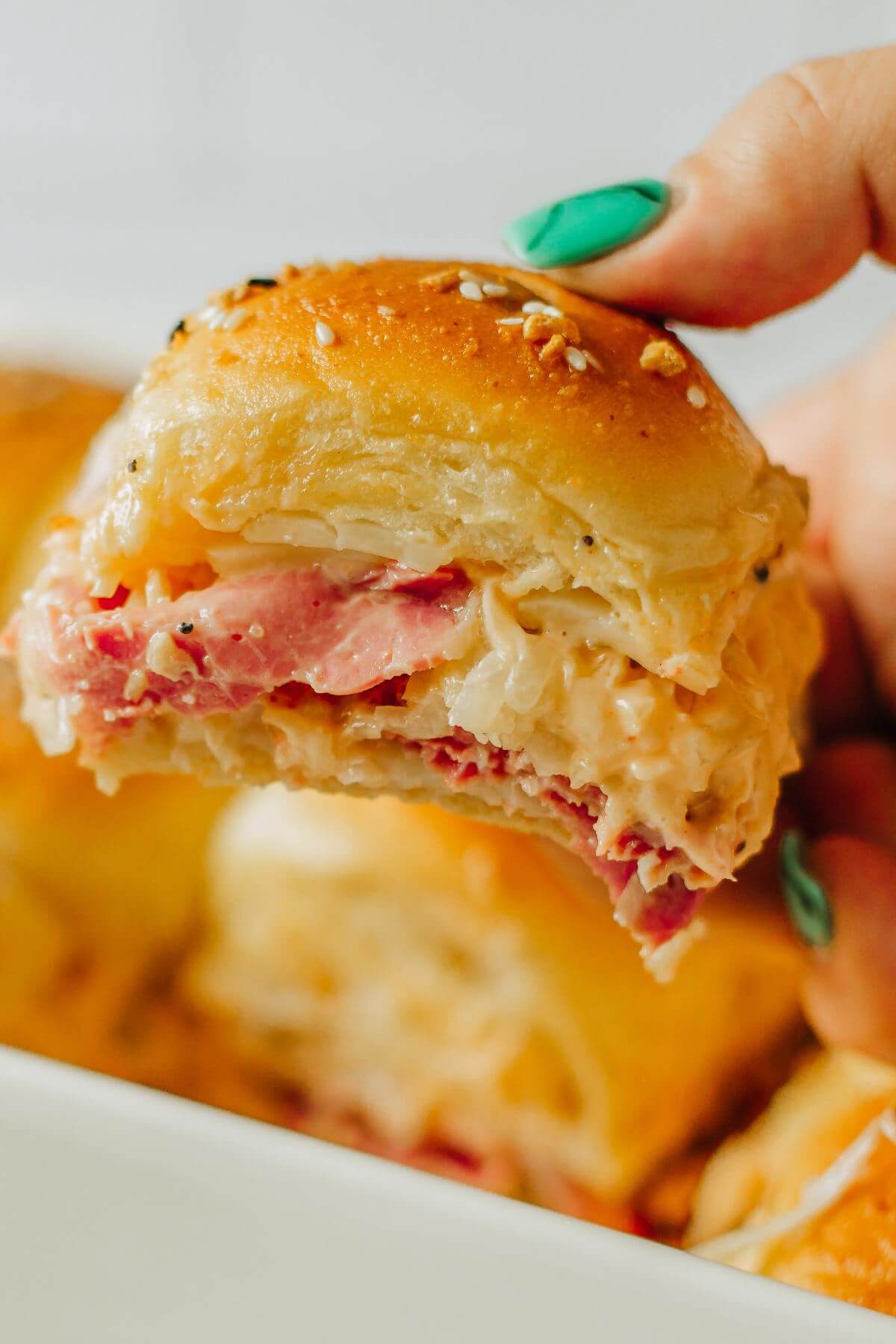 A hand holds up a slider showing meat and cheesy layers.