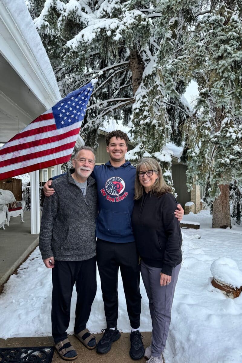 Three people standing outside for a picture with snow on the ground and a flag in background. 