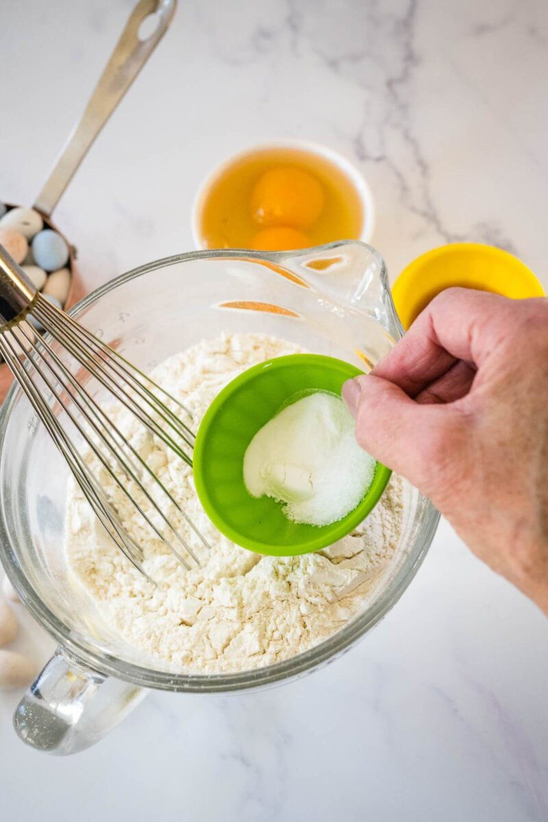A hand holds a small cup of white dry ingredients above a mixing bowl full of flour.
