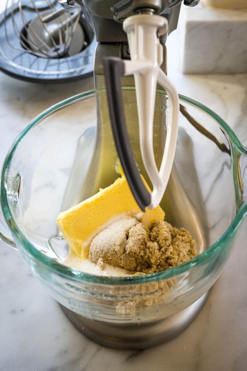 The bowl of a stand mixer has a block of butter and pile of sugar inside.
