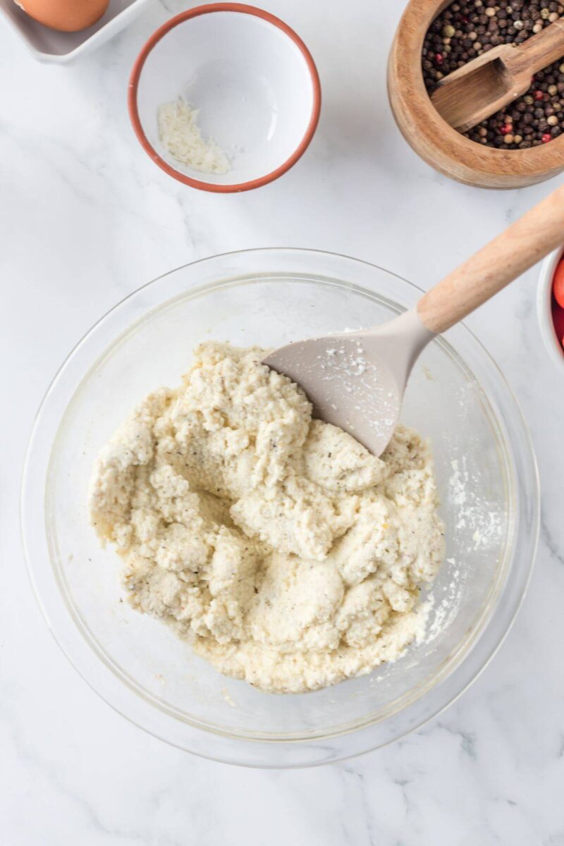 A spatula stirs ricotta cheese in a mixing bowl with spices.