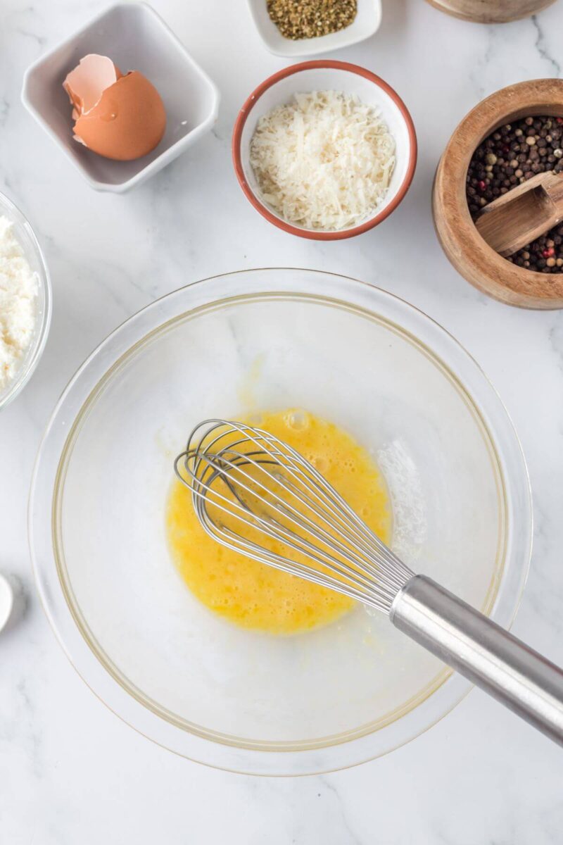 A whisk stirs egg in a mixing bowl near other ingredients.
