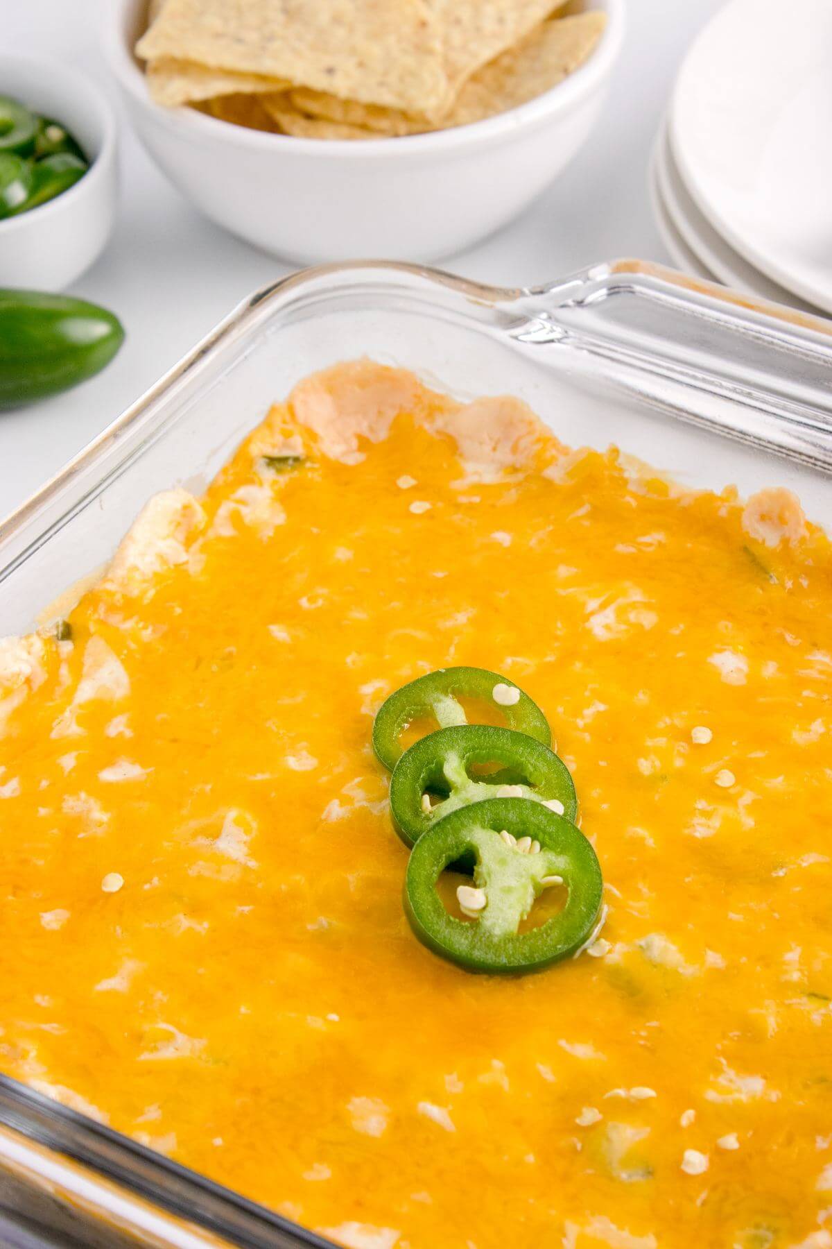 A glass casserole dish full of cheese and jalapenos sits next to tortilla chips and more jalapenos.