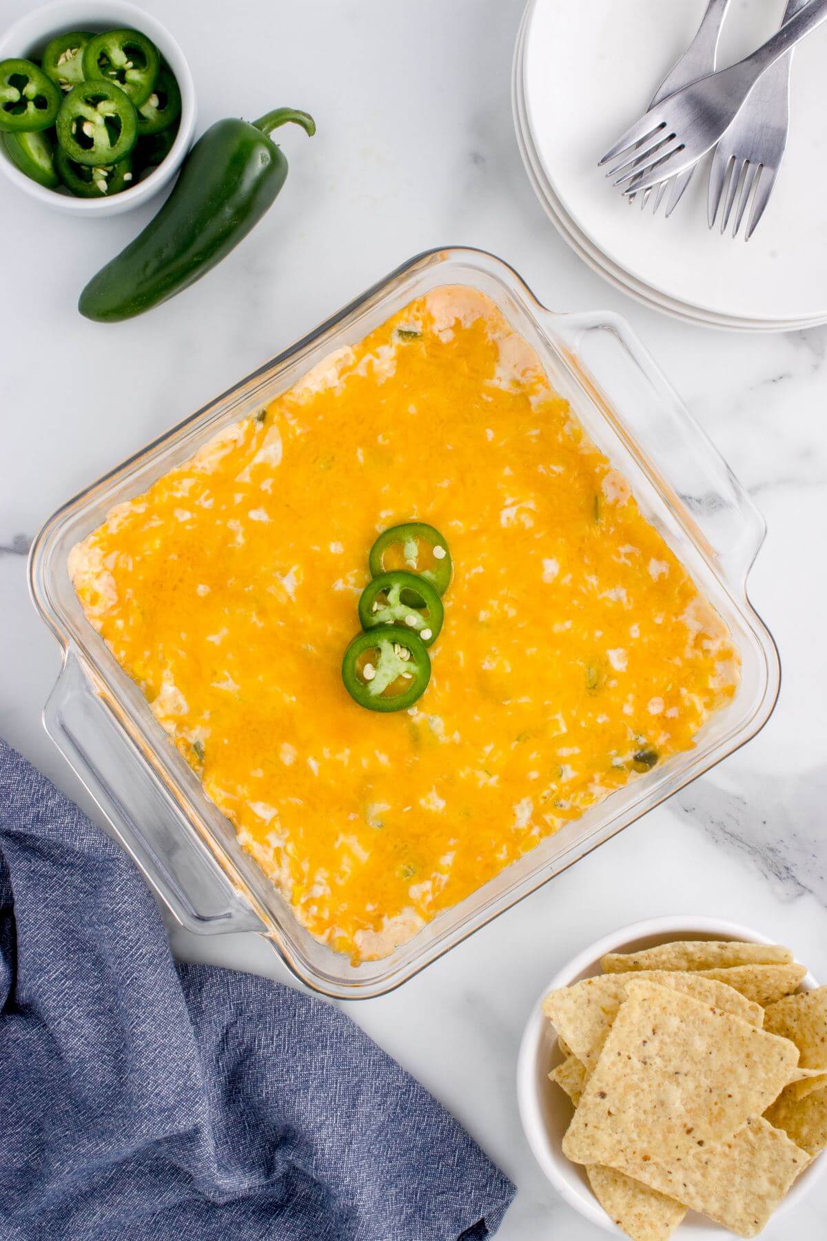A square glass pan is filled with yellow cheese topped casserole near jalapeno peppers and tortilla chips.