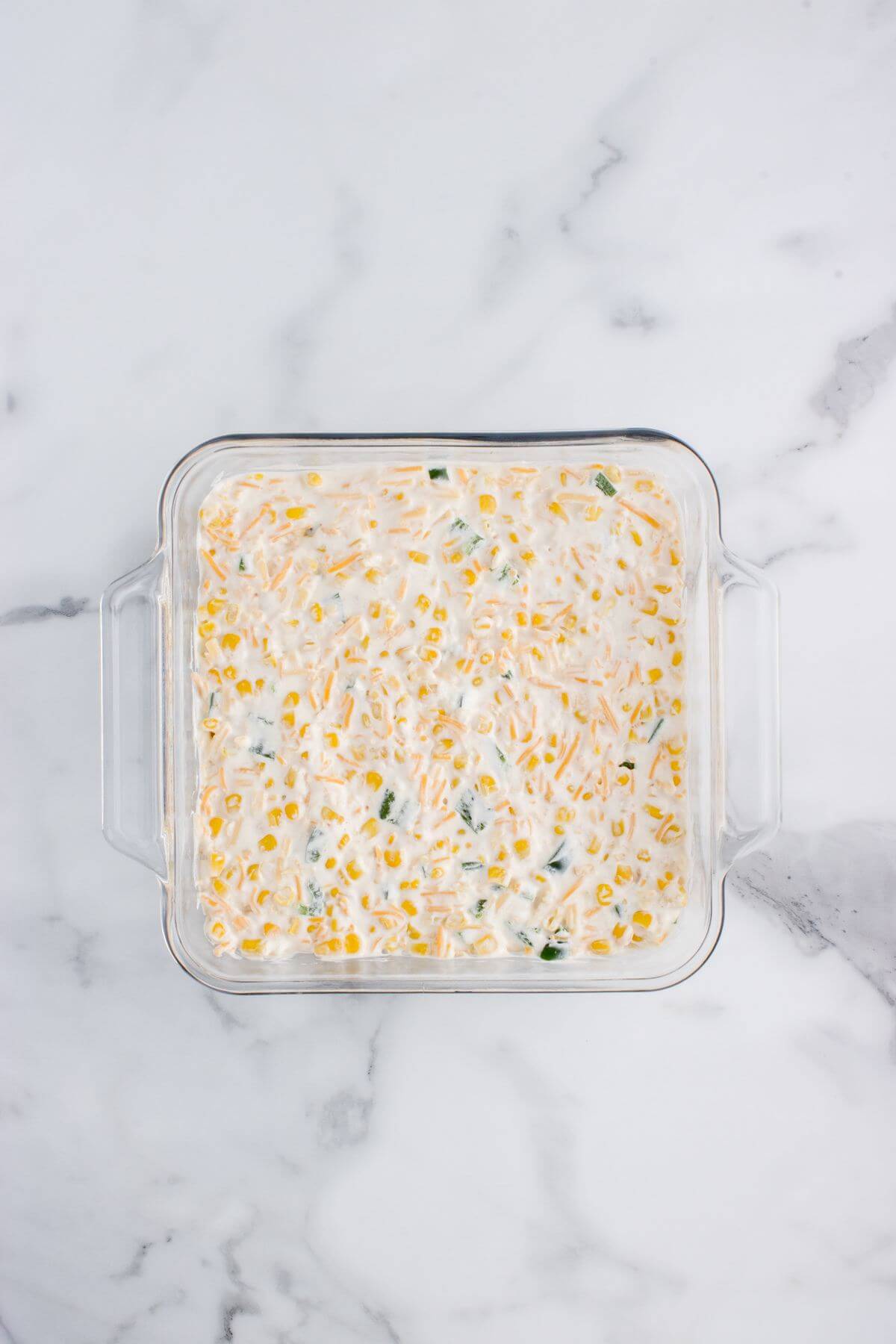 A glass square pan is filled with white creamy casserole mixture showing corn kernels.