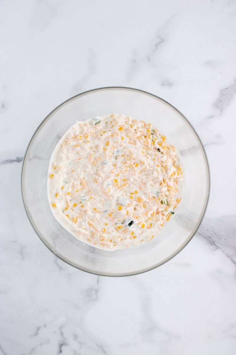 A glass mixing bowl holds melted cream cheese, corn kernels, and jalapeno pieces.