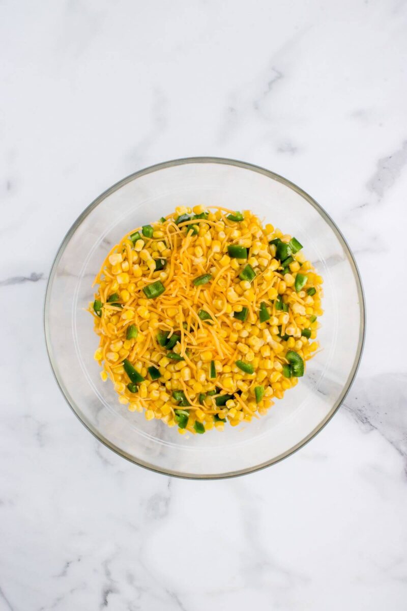Corn, diced jalapeno, and shredded cheese are mixed in a glass bowl.