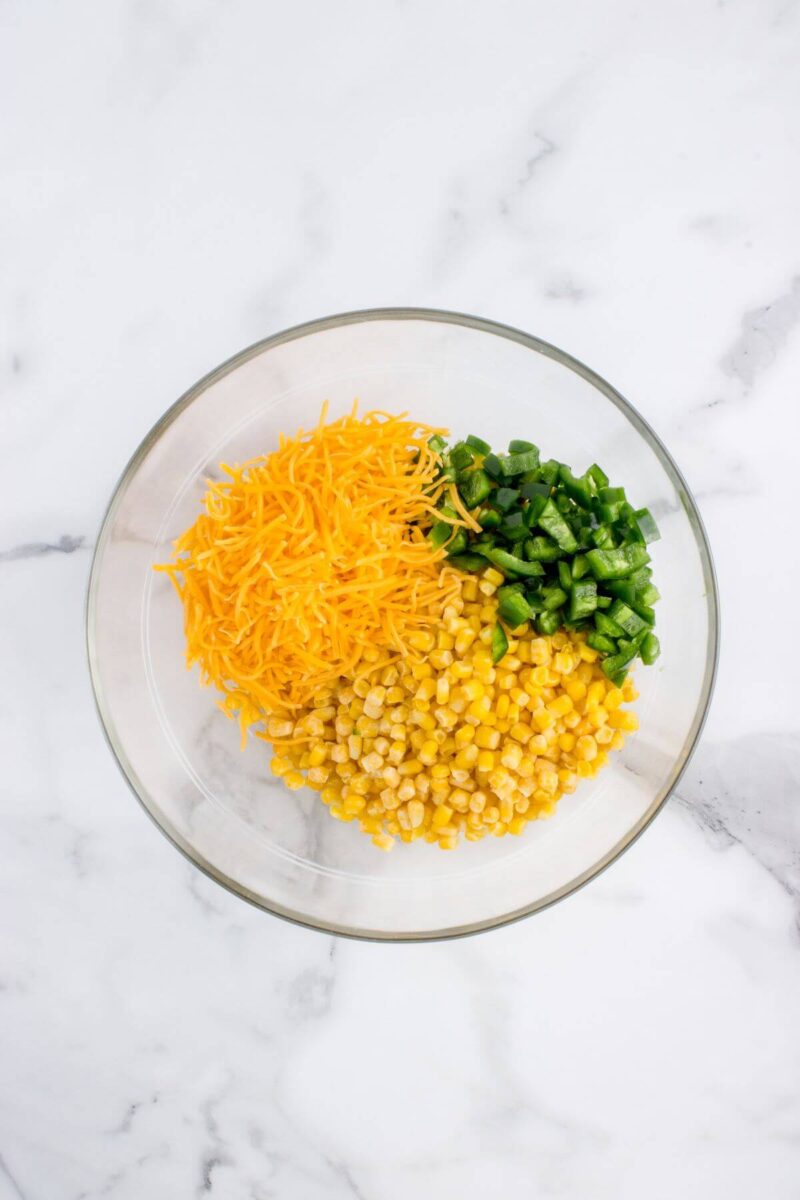 Corn, shredded cheddar cheese, and jalapeno pieces are in sections in a glass bowl.