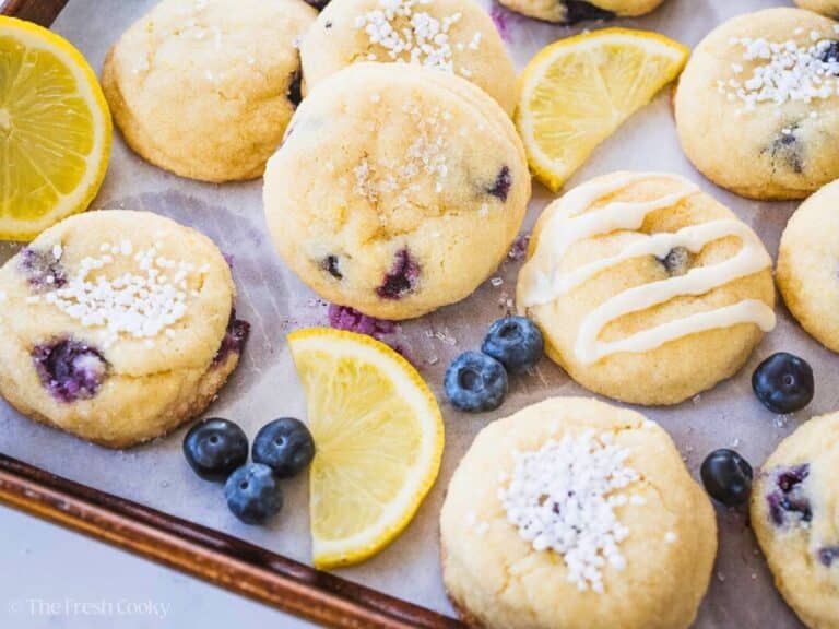 On a baking tray lined with parchment paper, blueberry lemon cookies topped with sugars, and drizzles, surrounded by fresh blueberries and lemon slices.