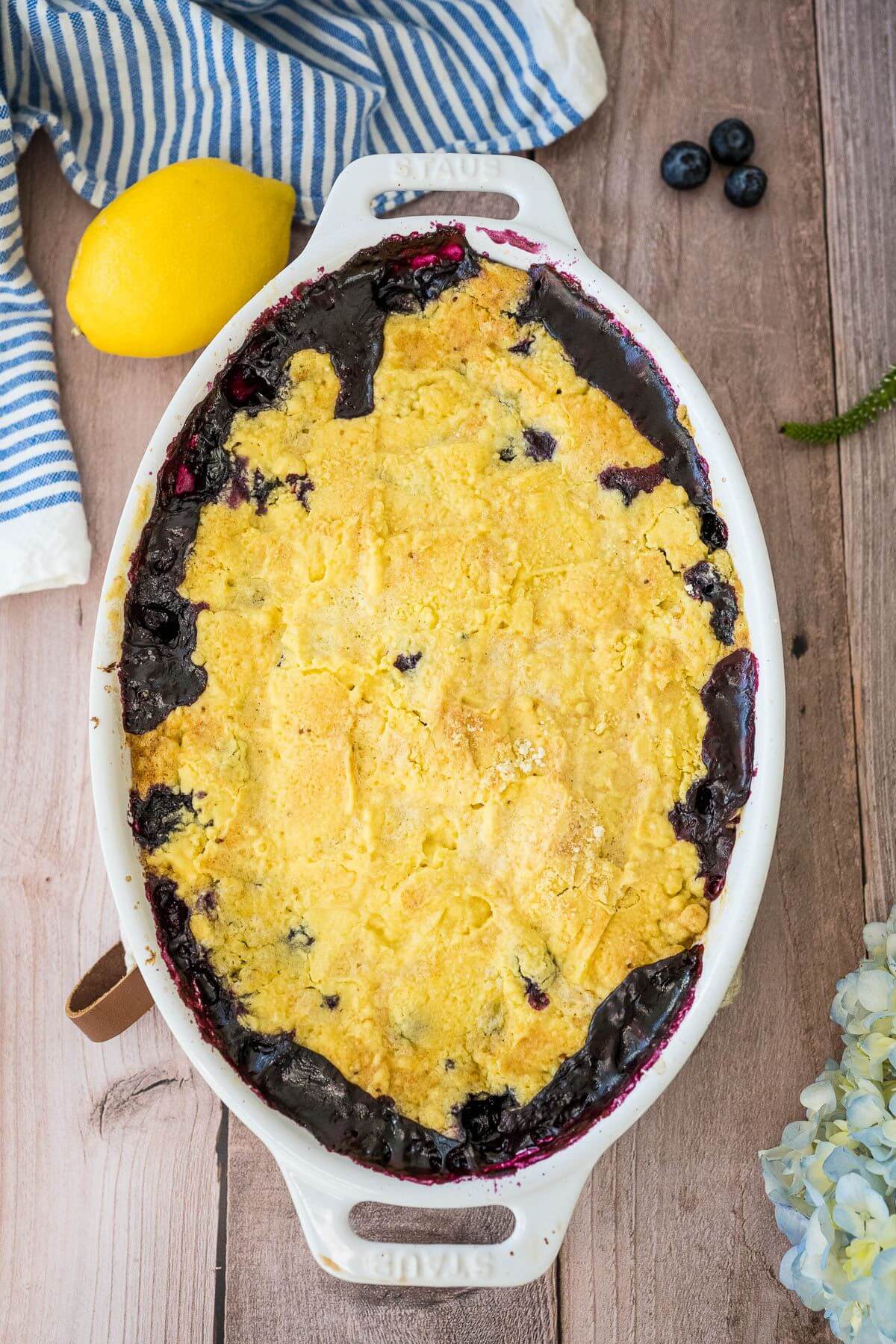 A baking dish full of blueberry and lemon cake rests next to more fruit, flowers, and a fine cloth.