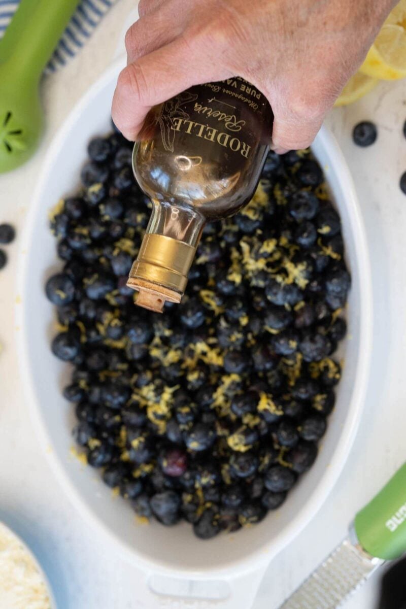 A hand pours out vanilla extract from a bottle on top of the blueberries in baking dish.