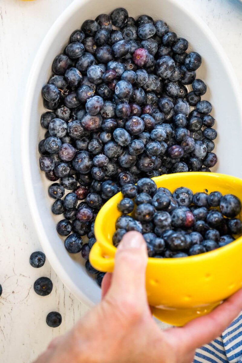 A hand pours fresh blueberries from a yellow cup into a baking dish.