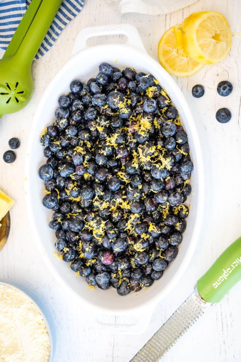 Yellow lemon zest is sprinkled over blueberries in a baking dish next to fruit and a knife.