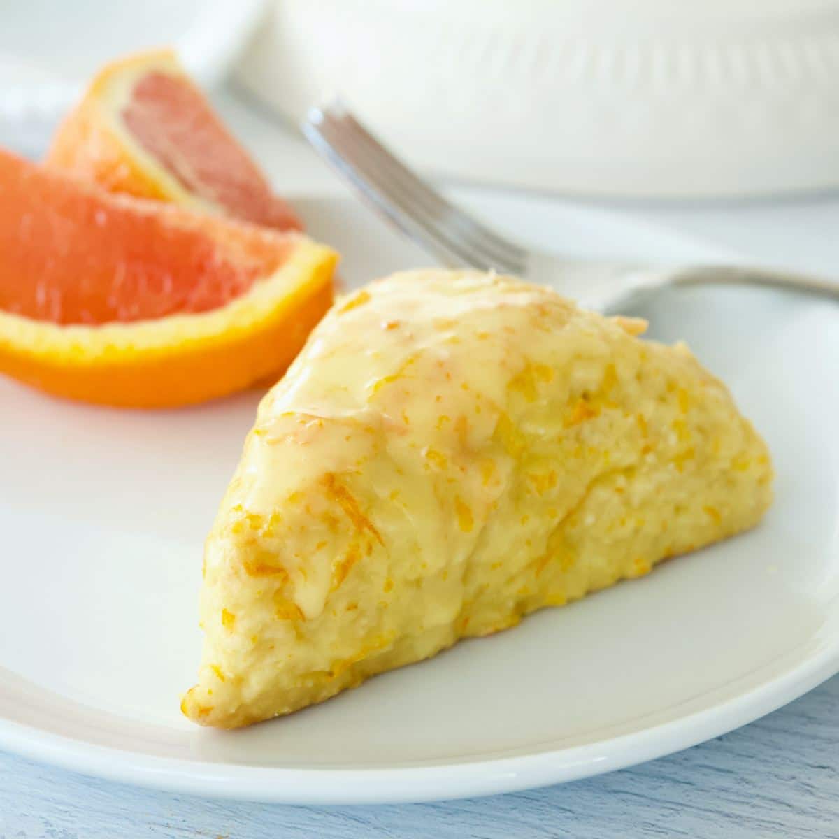 Single orange scone on a white plate with sections of cut orange behind.