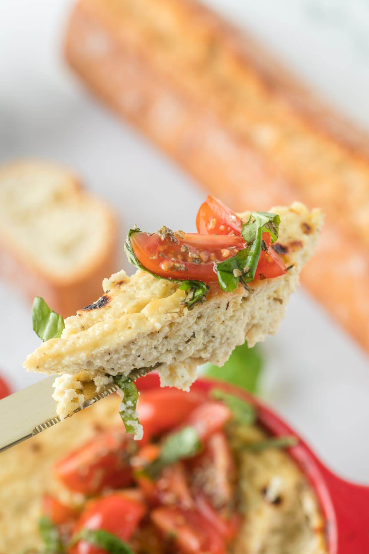 A bite of baked cheese dip and tomato herb mix sits on the end of a knife over the dish.
