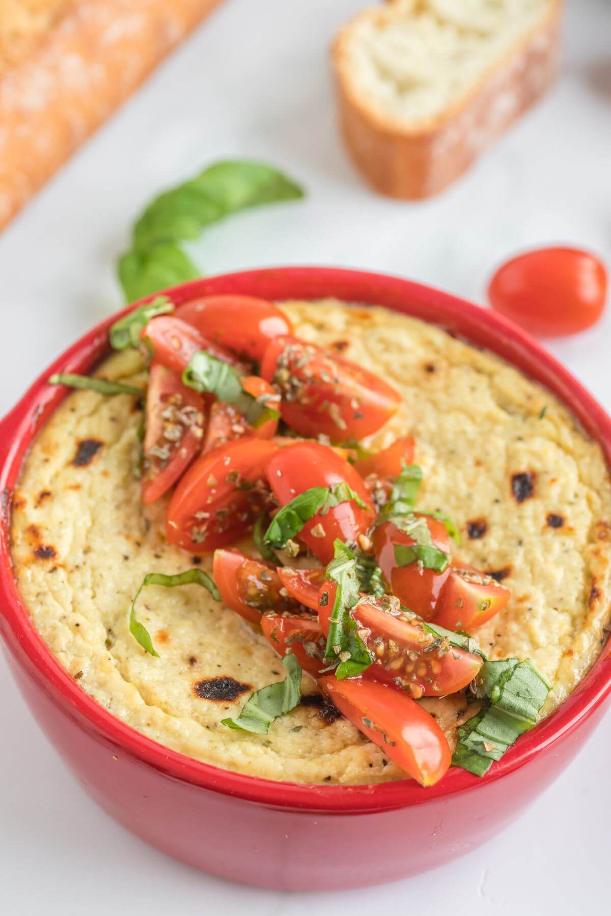 Tomatoes and herbs top a red ramekin full of browned cheese dip.