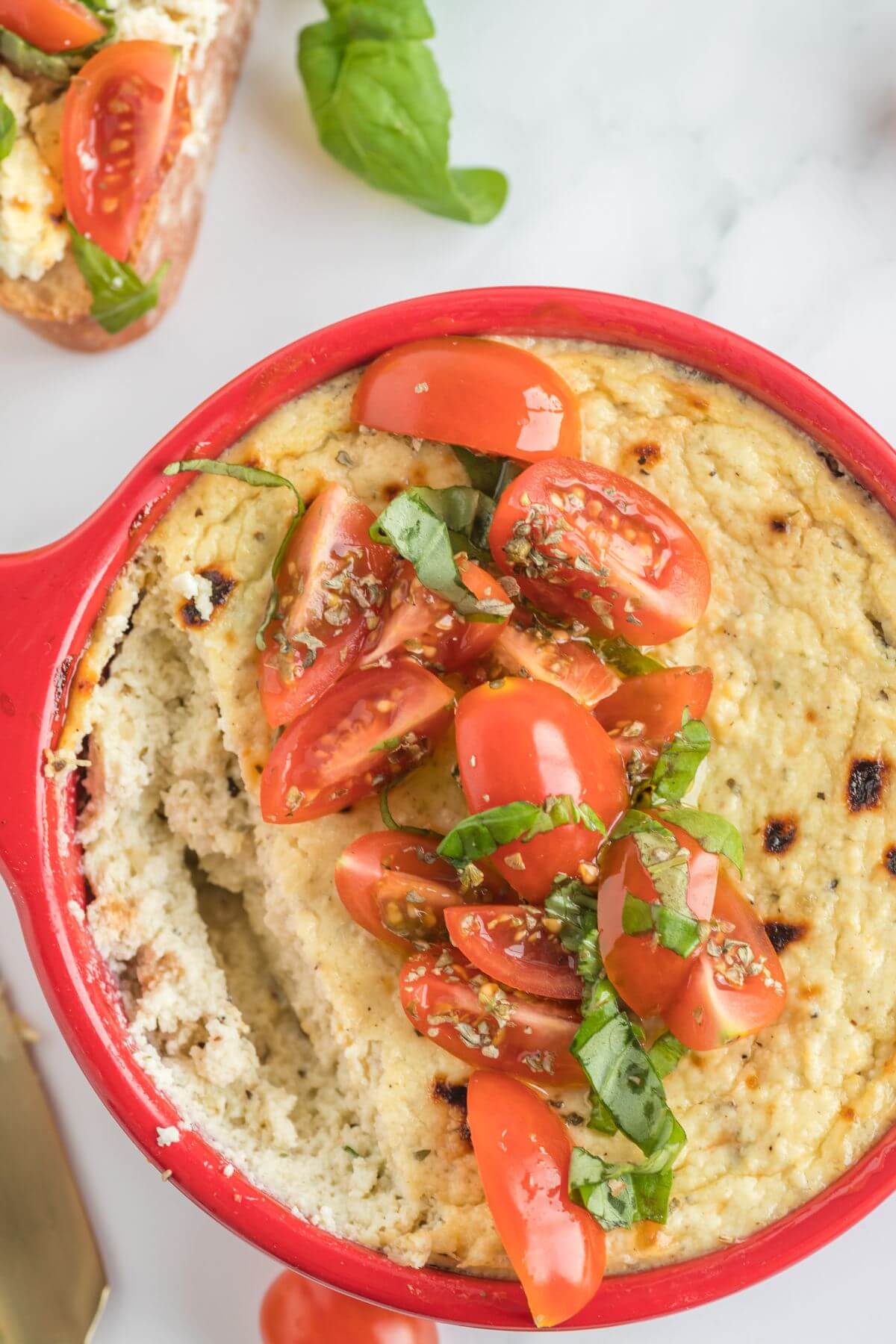 Cherry tomato slices and basil shreds are on top of cheese dip in a red dish.