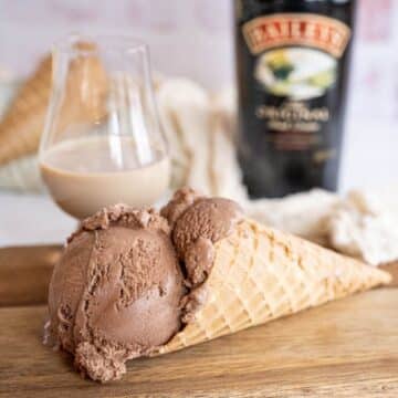 Two scoops of creamy chocolate and Bailey's Irish Cream Ice Cream in a waffle cone, with a snifter of Bailey's behind as well as a bottle.