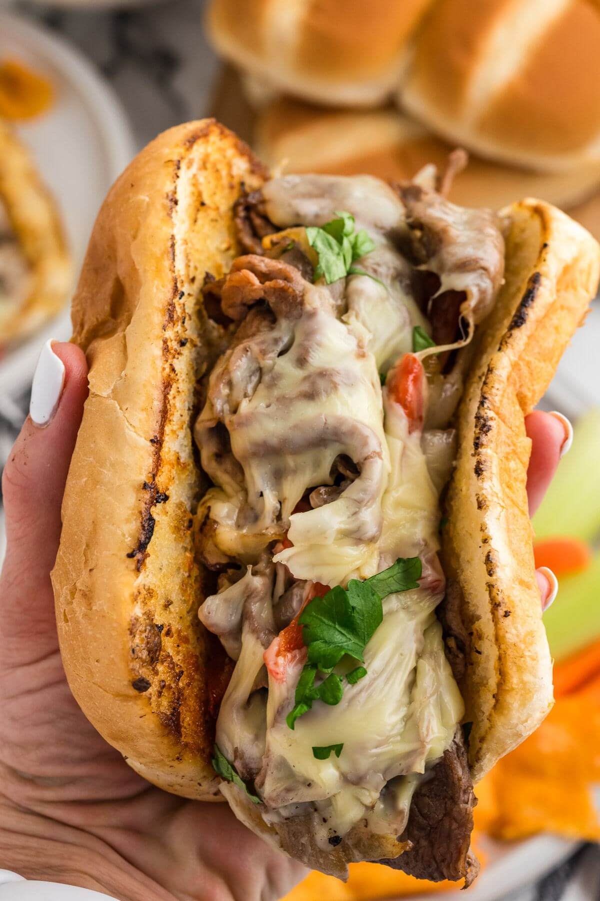 A hand holds a thick, gooey Philly cheesesteak sandwich.