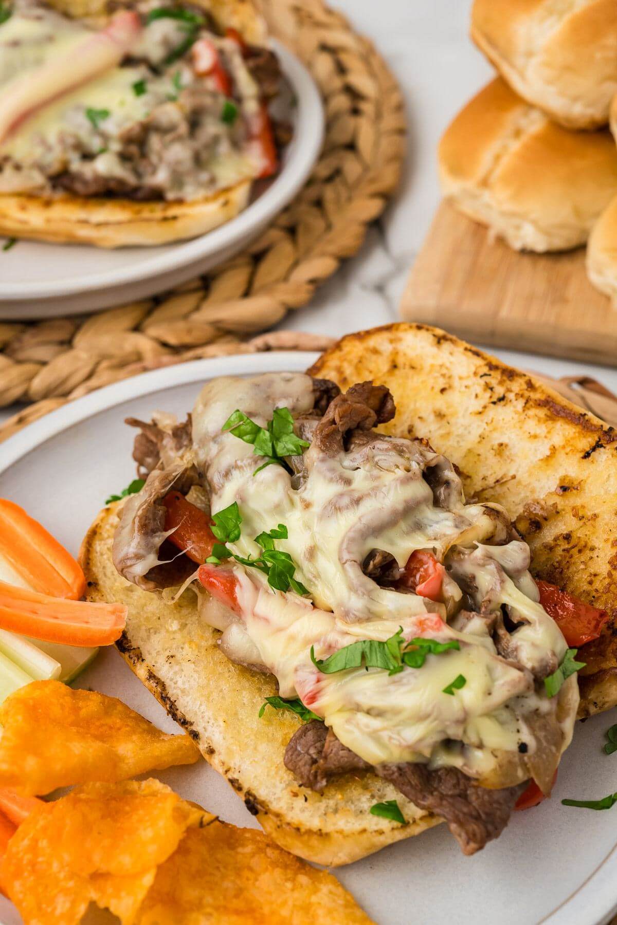 Potato chips, carrots, and more Hoagie rolls sit next to open filled cheesesteak sandwiches on plates.