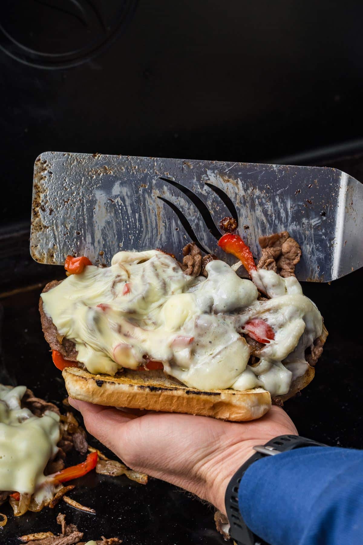 Hand holding a Philly Cheesesteak over a Blackstone griddle with spatula placing meat and cheese mixture inside.