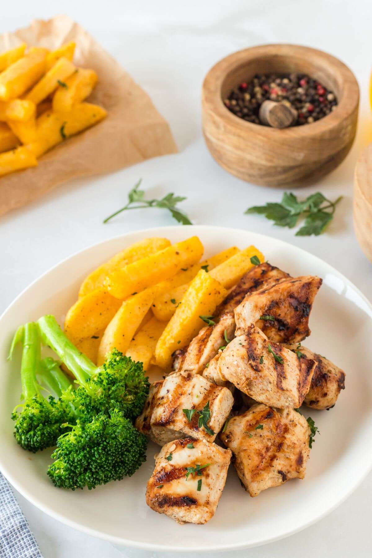 A plate of grilled nuggets, broccoli, and French fries sits next to more fries and black pepper.