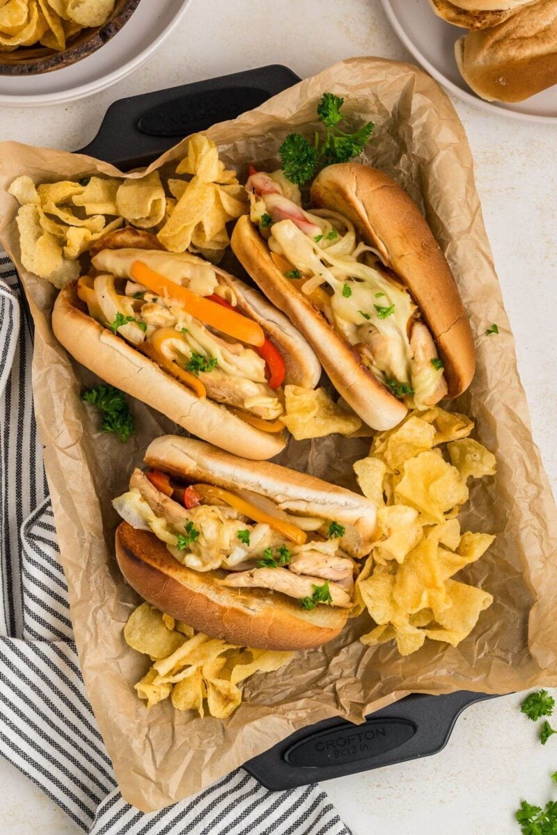 A pan with parchment paper is covered in potato chips and three thick filled cheesesteaks with peppers.