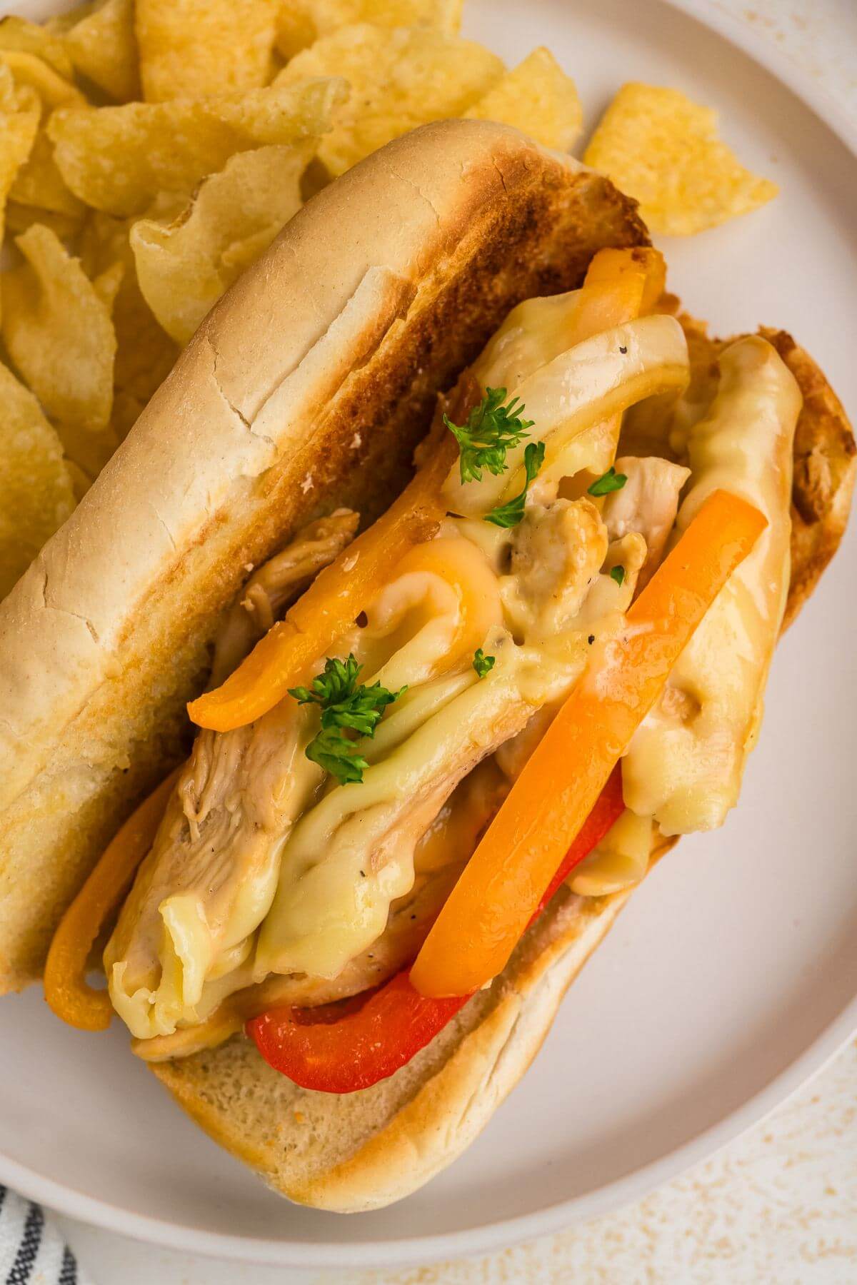 Gooey cheese, tender chicken chunks, and bright bell peppers fill a Hoagie roll next to potato chips.