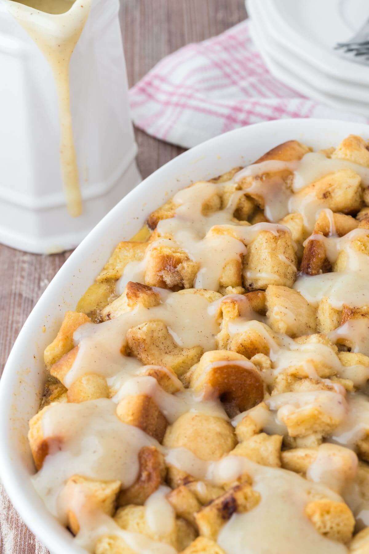 A completely full baking dish shows morsels of cinnamon roll mixed together with a white glaze next to a pitcher of glaze.