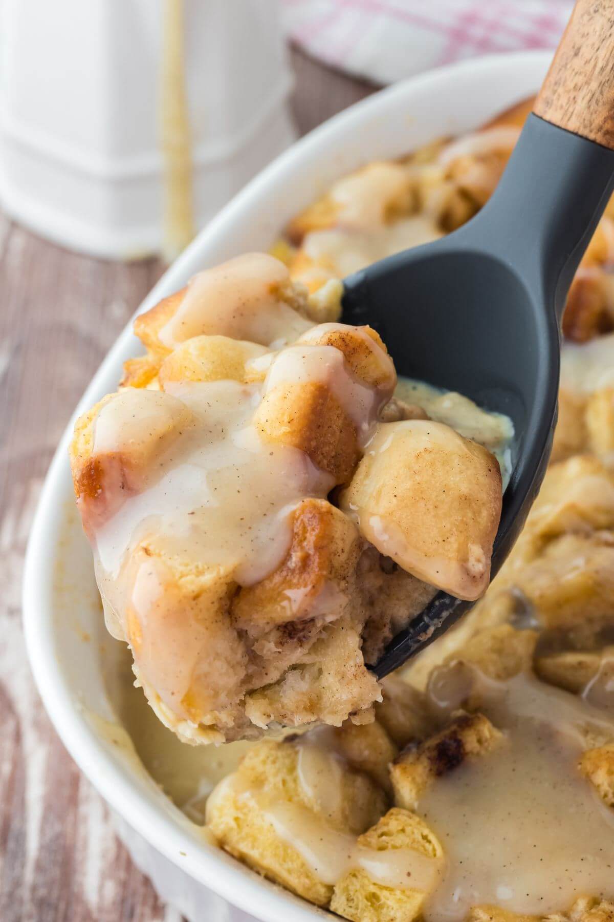 A dark serving spoon scoops up gooey bread pudding chunks above the full baking dish.