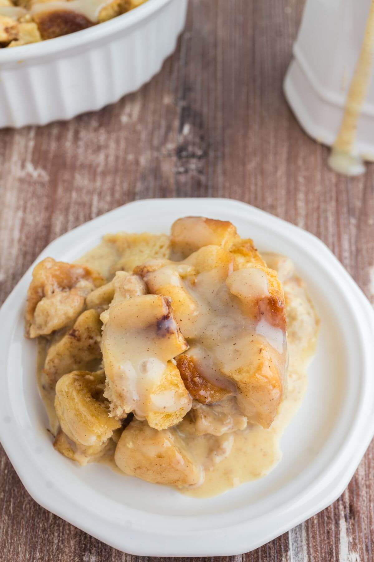 Translucent white glaze coats gooey cinnamon roll chunks in pudding served on a white plate.